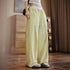 Person wearing light green wide-leg pants in a room with wooden flooring and a curtain.